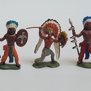 May include: Three painted metal toy figurines of Native American warriors. Each figure wears a feathered headdress and carries a spear and a shield. The figures are depicted in dynamic poses, featuring detailed clothing in orange, red, and grey colors.