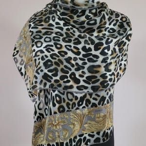 Puede incluir: Un pañuelo ligero con un estampado de leopardo en negro, blanco y beige. El pañuelo incluye un borde ancho con fondo gris y un diseño de hojas de acanto doradas. La palabra Loredano está impresa en letras pequeñas sobre el borde del tejido.