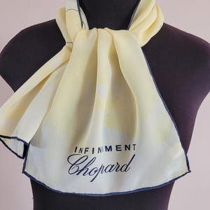 May include: A pale yellow scarf with a navy blue border and the text "INFINIMENT Chopard." The scarf is draped over a mannequin. The scarf has a light, airy appearance.