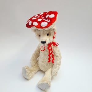 May include: A cream-colored teddy bear wearing a red mushroom cap with white spots and a red ribbon around its neck. The plush toy is sitting with its legs extended and its arms at its sides. The bear has a black nose and small, dark eyes.