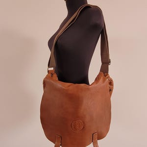 May include: A tan leather crossbody bag featuring a rounded flap, a wide adjustable fabric shoulder strap, and two vertical leather tabs at the bottom. A circular embossed logo is centered on the front flap of this soft-structured leather bag.