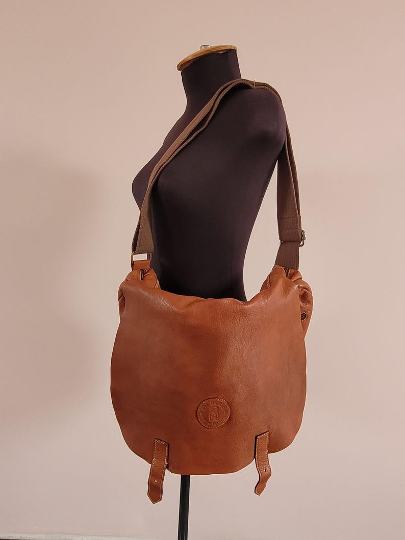 May include: A tan leather crossbody bag featuring a rounded flap, a wide adjustable fabric shoulder strap, and two vertical leather tabs at the bottom. A circular embossed logo is centered on the front flap of this soft-structured leather bag.