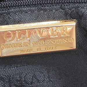 May include: A rectangular gold-toned metal plaque attached to a black fabric surface. The plaque features embossed text that reads OLIVER, MADE IN VALENTINO, and MADE IN ITALY. The metal surface is reflective and smooth with a polished finish.