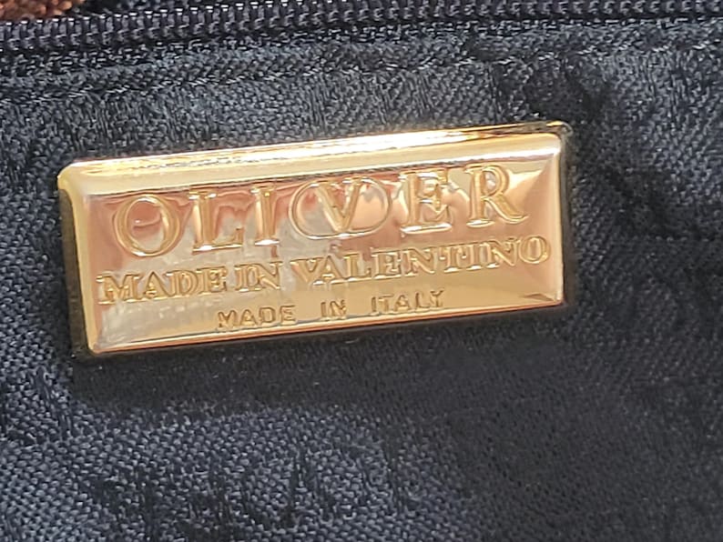 May include: A rectangular gold-toned metal plaque attached to a black fabric surface. The plaque features embossed text that reads OLIVER, MADE IN VALENTINO, and MADE IN ITALY. The metal surface is reflective and smooth with a polished finish.
