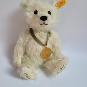 May include: A cream-colored teddy bear with a gold medal around its neck. The bear has black eyes and a black nose. The bear's foot has the name "Aria" embroidered in green. A yellow tag with text is attached to the bear's ear.