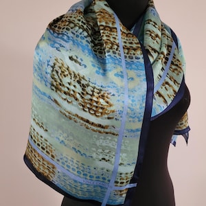 May include: A square scarf featuring an abstract pattern in shades of light blue, teal, and brown with a dark navy blue border. The scarf displays a repeating textured design and includes the text VINCE CAMUTO printed in the lower corner area.