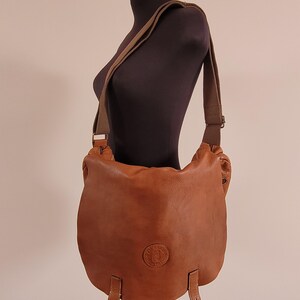 May include: A tan leather messenger bag with a large front flap, two vertical leather straps at the bottom, and a wide adjustable fabric shoulder strap. The bag features a circular embossed logo on the front center and metal hardware accents.