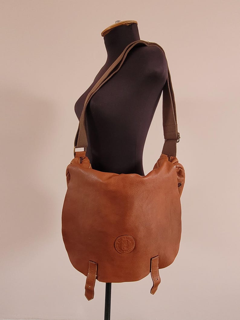 May include: A tan leather messenger bag with a large front flap, two vertical leather straps at the bottom, and a wide adjustable fabric shoulder strap. The bag features a circular embossed logo on the front center and metal hardware accents.