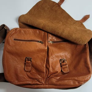 May include: A tan leather messenger bag featuring a large fold-over flap, two front pockets with brass-toned buckle closures, two horizontal zippered compartments, and a wide brown fabric shoulder strap with matching leather accents.