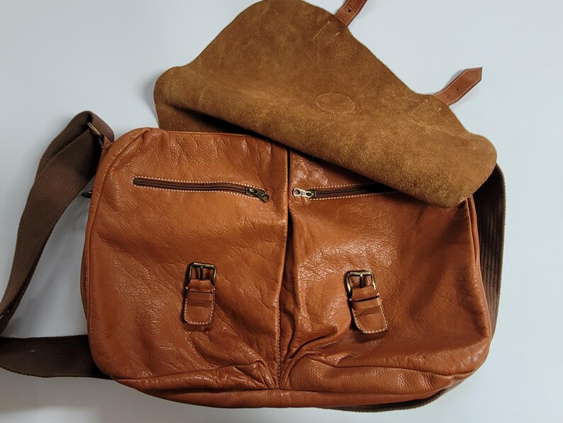 May include: A tan leather messenger bag featuring a large fold-over flap, two front pockets with brass-toned buckle closures, two horizontal zippered compartments, and a wide brown fabric shoulder strap with matching leather accents.