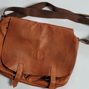 May include: A tan leather messenger bag featuring a large front flap with two vertical leather straps and a circular embossed logo in the center. The bag includes a wide, adjustable brown fabric shoulder strap attached with metal hardware.