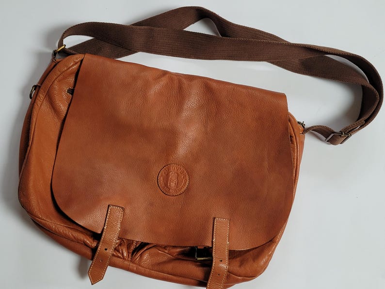 May include: A tan leather messenger bag featuring a large front flap with two vertical leather straps and a circular embossed logo in the center. The bag includes a wide, adjustable brown fabric shoulder strap attached with metal hardware.