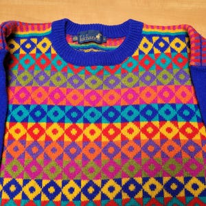 May include: A crew neck sweater with a vibrant geometric pattern in red, yellow, blue, green, and purple. The sleeves and collar are solid blue. The sweater has a tag that reads "Tukhan".