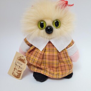 May include: A handmade plush toy with a fluffy, light-yellow head, large green eyes, and a black nose. It wears a plaid dress with a white collar and has a red bow. A tag reads "Rosie Mitten".