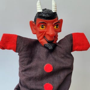 Könnte beinhalten: Eine Teufel-Handpuppe mit rotem Gesicht, gelben Augen und weißen Hörnern. Die Puppe hat einen schwarzen Bart und Haare und trägt eine braune Fischgrätjacke mit roten Ärmeln und zwei roten Knöpfen.
