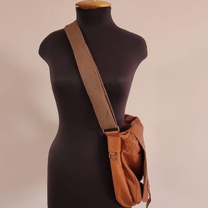 May include: A tan leather messenger bag with a wide, textured brown fabric shoulder strap. The bag features a fold-over flap, visible stitching, and antique brass-toned metal hardware, including a D-ring attachment on the side of the bag.