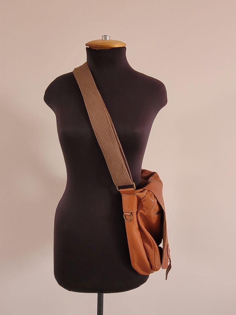 May include: A tan leather messenger bag with a wide, textured brown fabric shoulder strap. The bag features a fold-over flap, visible stitching, and antique brass-toned metal hardware, including a D-ring attachment on the side of the bag.