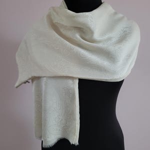 May include: An off-white scarf featuring a tonal, woven paisley pattern throughout the fabric. The scarf has a soft, draped appearance with a subtle sheen and a delicate, frayed edge finish at the bottom, displayed on a black mannequin form.