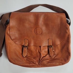May include: A tan leather messenger bag featuring a large front flap with two buckle straps and a circular embossed logo in the center. The bag includes a wide brown fabric shoulder strap and has a textured, soft appearance with visible folds.