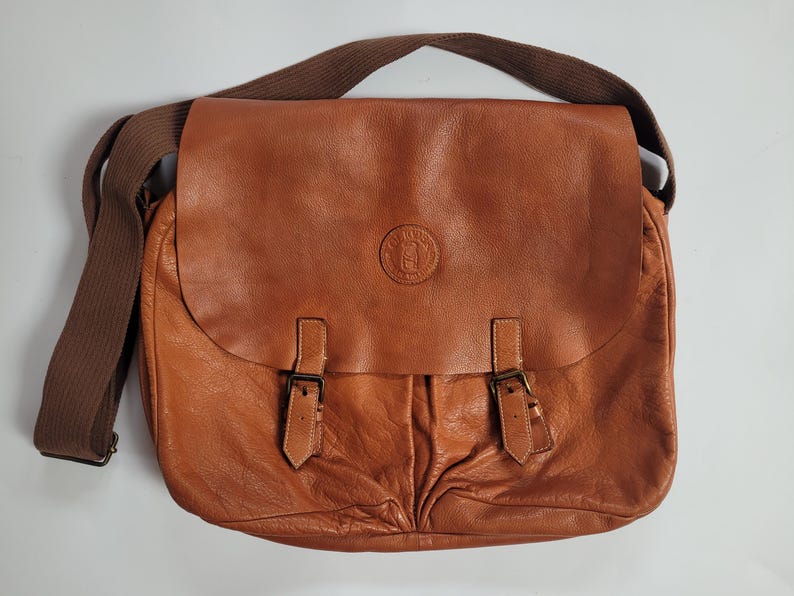 May include: A tan leather messenger bag featuring a large front flap with two buckle straps and a circular embossed logo in the center. The bag includes a wide brown fabric shoulder strap and has a textured, soft appearance with visible folds.