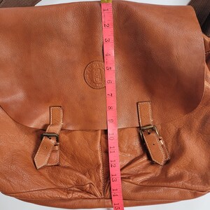 May include: A tan leather messenger bag with a fold-over flap, two buckle straps, and a brown fabric shoulder strap. A pink measuring tape is placed vertically over the center, showing a length of approximately 18 inches from top to bottom.