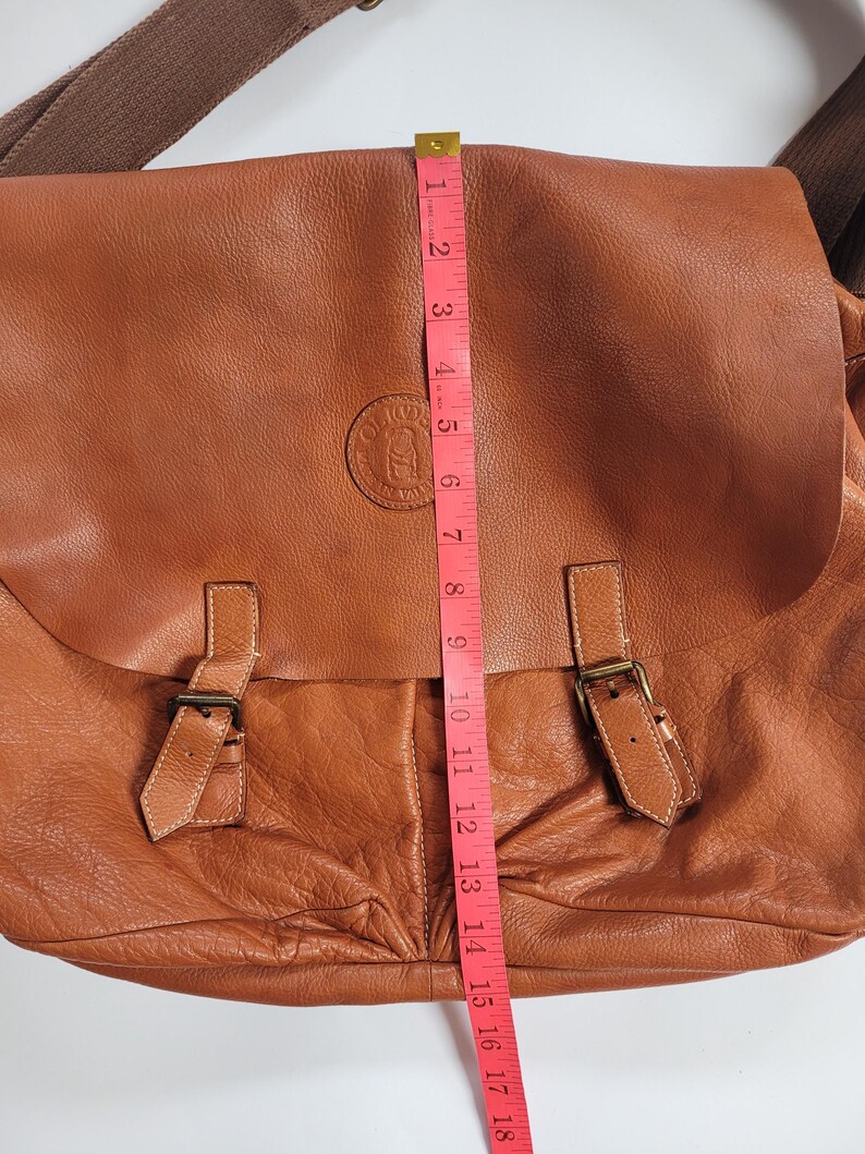 May include: A tan leather messenger bag with a fold-over flap, two buckle straps, and a brown fabric shoulder strap. A pink measuring tape is placed vertically over the center, showing a length of approximately 18 inches from top to bottom.