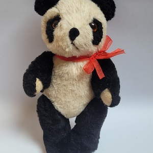 May include: A plush panda bear toy with cream-coloured fur on the head and torso, contrasted by black fur on the ears, limbs, and around the eyes. The panda features dark brown plastic eyes, a black stitched nose, and a bright red sheer ribbon bow.