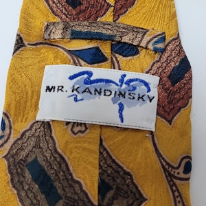 May include: A yellow necktie featuring a repeating abstract pattern in shades of brown and dark blue. A white fabric label is stitched onto the back, displaying the text MR. KANDINSKY in black lettering with a blue graphic design above the name.