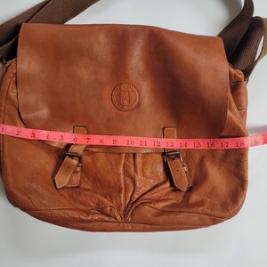 May include: A tan leather messenger bag with a brown fabric shoulder strap, a front flap featuring a circular embossed logo, and two buckle closures. A pink measuring tape is placed across the front, showing a width of approximately 19 inches.