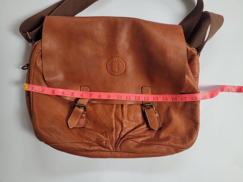 May include: A tan leather messenger bag with a brown fabric shoulder strap, a front flap featuring a circular embossed logo, and two buckle closures. A pink measuring tape is placed across the front, showing a width of approximately 19 inches.
