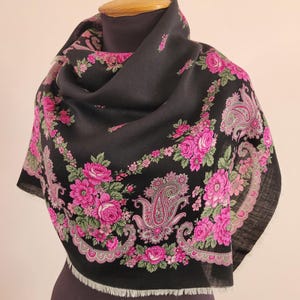 May include: A black shawl with a floral design featuring pink roses and paisley patterns. The shawl has a fringed edge and is draped over a mannequin. The floral pattern is framed by a pink border.
