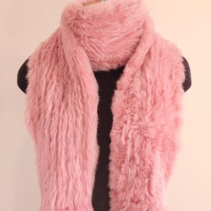 May include: A fluffy, pink scarf made of soft material. The scarf is draped over a black mannequin, showcasing its texture and color. The scarf appears to be warm and cozy, suitable for cold weather.