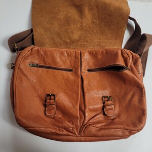 May include: A tan leather messenger bag featuring a fold-over flap, two front pockets with zipper closures, and two decorative buckle straps. The bag includes a brown fabric shoulder strap and displays a textured, natural grain finish throughout.