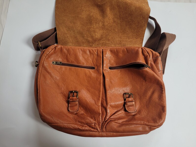 May include: A tan leather messenger bag featuring a fold-over flap, two front pockets with zipper closures, and two decorative buckle straps. The bag includes a brown fabric shoulder strap and displays a textured, natural grain finish throughout.