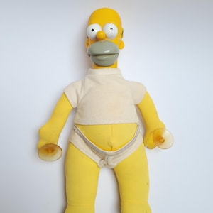 May include: A yellow character doll with a bald head, large white eyes, and a grey mouth area. The doll wears a short-sleeved cream-coloured shirt and cream-coloured underwear. Each hand features a clear plastic suction cup attached to the palm.