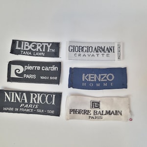 Puede incluir: Colección de etiquetas de ropa de diseñador, incluyendo Liberty, Pierre Cardin, Nina Ricci, Giorgio Armani, Kenzo y Pierre Balmain. Las etiquetas son de varios colores, con texto en blanco o negro.
