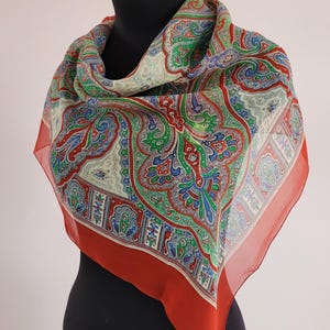 May include: A colorful paisley patterned scarf with a red border is draped over a mannequin. The scarf features a cream-colored background with intricate designs in red, green, and blue. The scarf is made of a lightweight material.