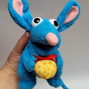 May include: A blue plush rat toy with large eyes and ears. The rat has a pink nose and ear linings. It holds a yellow cheese-shaped pouch tied with a red ribbon. The toy is made of soft, textured fabric.