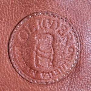 May include: A textured tan leather surface featuring a circular embossed logo. The circular patch is stitched with light-colored thread and displays a central bulldog graphic surrounded by the text OLIVIERA and SAN VALENTINO in a circular pattern.