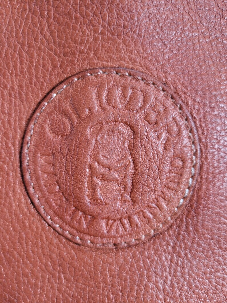 May include: A textured tan leather surface featuring a circular embossed logo. The circular patch is stitched with light-colored thread and displays a central bulldog graphic surrounded by the text OLIVIERA and SAN VALENTINO in a circular pattern.