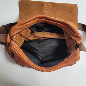 May include: A tan leather messenger bag with a fold-over flap and a brown fabric shoulder strap. The interior is lined with black fabric featuring the repeated text Oliver Jecker. The bag has a zippered main compartment and visible leather texture.