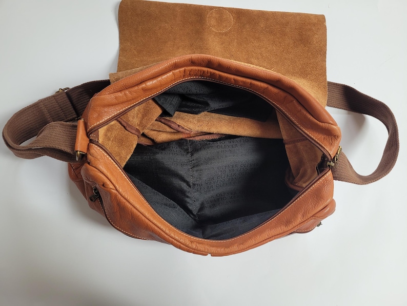 May include: A tan leather messenger bag with a fold-over flap and a brown fabric shoulder strap. The interior is lined with black fabric featuring the repeated text Oliver Jecker. The bag has a zippered main compartment and visible leather texture.