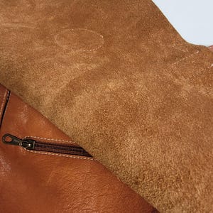 May include: A tan leather bag featuring a smooth, polished exterior surface and a contrasting interior flap made of textured, unfinished suede. A horizontal zipper with a dark metal pull is integrated into the main body of the bag, positioned just below the flap.