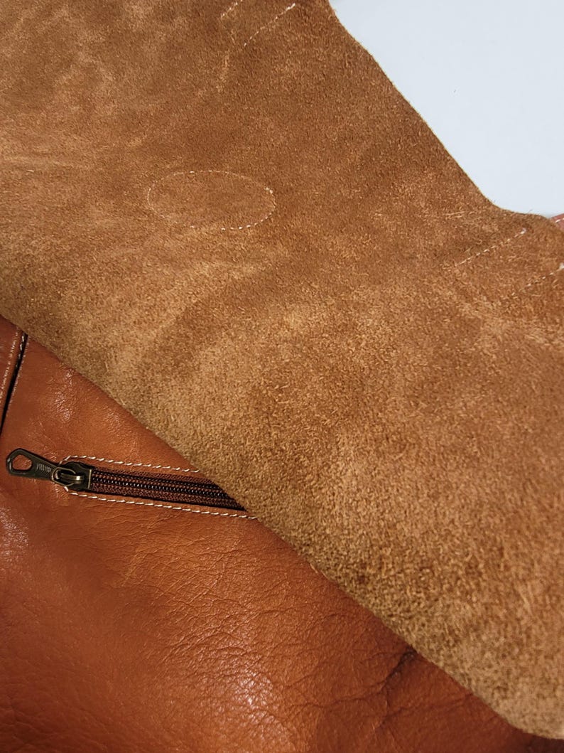 May include: A tan leather bag featuring a smooth, polished exterior surface and a contrasting interior flap made of textured, unfinished suede. A horizontal zipper with a dark metal pull is integrated into the main body of the bag, positioned just below the flap.