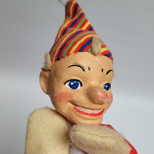 Puede incluir: Un títere con rostro pintado que presenta ojos azules, una amplia sonrisa y mejillas sonrosadas. Lleva un gorro puntiagudo con rayas multicolores en rojo, azul, amarillo y verde, mangas de fieltro beige y un cuerpo de tela roja con borde blanco.