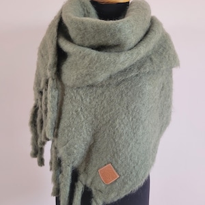 May include: A sage green oversized scarf with a fuzzy, brushed texture and long fringe detailing along the edges. The scarf features a small, diamond-shaped tan leather patch stitched onto the lower corner, displaying an embossed logo design.