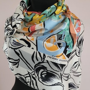 Pode incluir: Um lenço de seda com um design abstrato colorido, com tons de azul, amarelo, laranja e verde. O lenço tem um fundo branco com detalhes florais pretos e o texto "PIERRE BALMAIN".