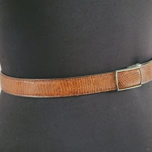 May include: A brown belt with a textured, reptile-like pattern. The belt is fastened with a silver-colored rectangular buckle. The belt is displayed on a black mannequin.