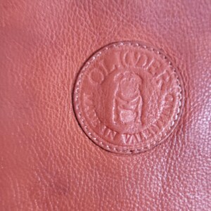 May include: A textured terracotta-colored leather surface featuring a circular embossed logo. The logo depicts a stylized bulldog at the center, surrounded by the text "OLIDER" at the top and "MADE IN VALENCIA" along the bottom edge.