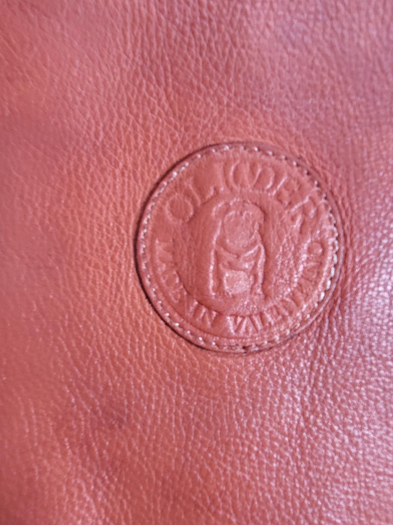 May include: A textured terracotta-colored leather surface featuring a circular embossed logo. The logo depicts a stylized bulldog at the center, surrounded by the text "OLIDER" at the top and "MADE IN VALENCIA" along the bottom edge.